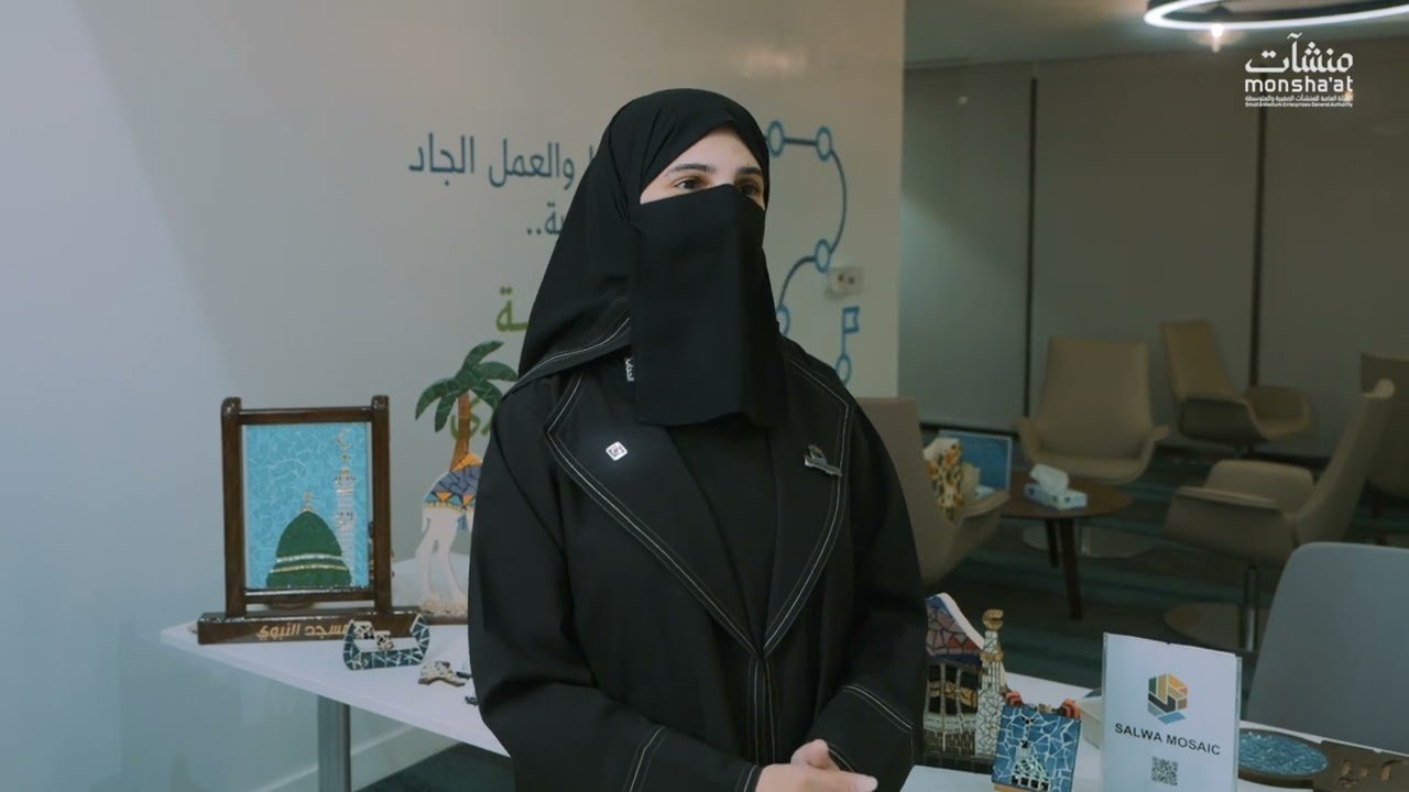 سلوى العمودي تتحدث في مقابلة مصورة مع منشآت، محاطة بلوحات فسيفساء للمسجد النبوي ومجسمات تراثية