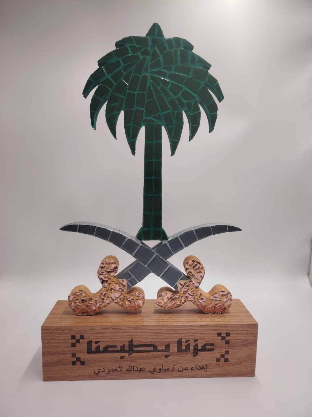 منحوتة فسيفساء ثلاثية الأبعاد لشعار المملكة العربية السعودية — نخلة وسيفان متقاطعان على قاعدة خشبية منقوش عليها عبارة «عزّنا بطبيعتنا» وإهداء من سلوى عبدالله العمودي
